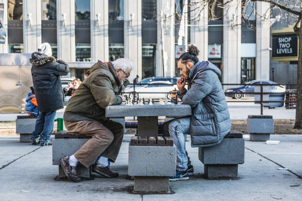 Two men playing chest in the middle of a city park with people and buildings surrounding them.