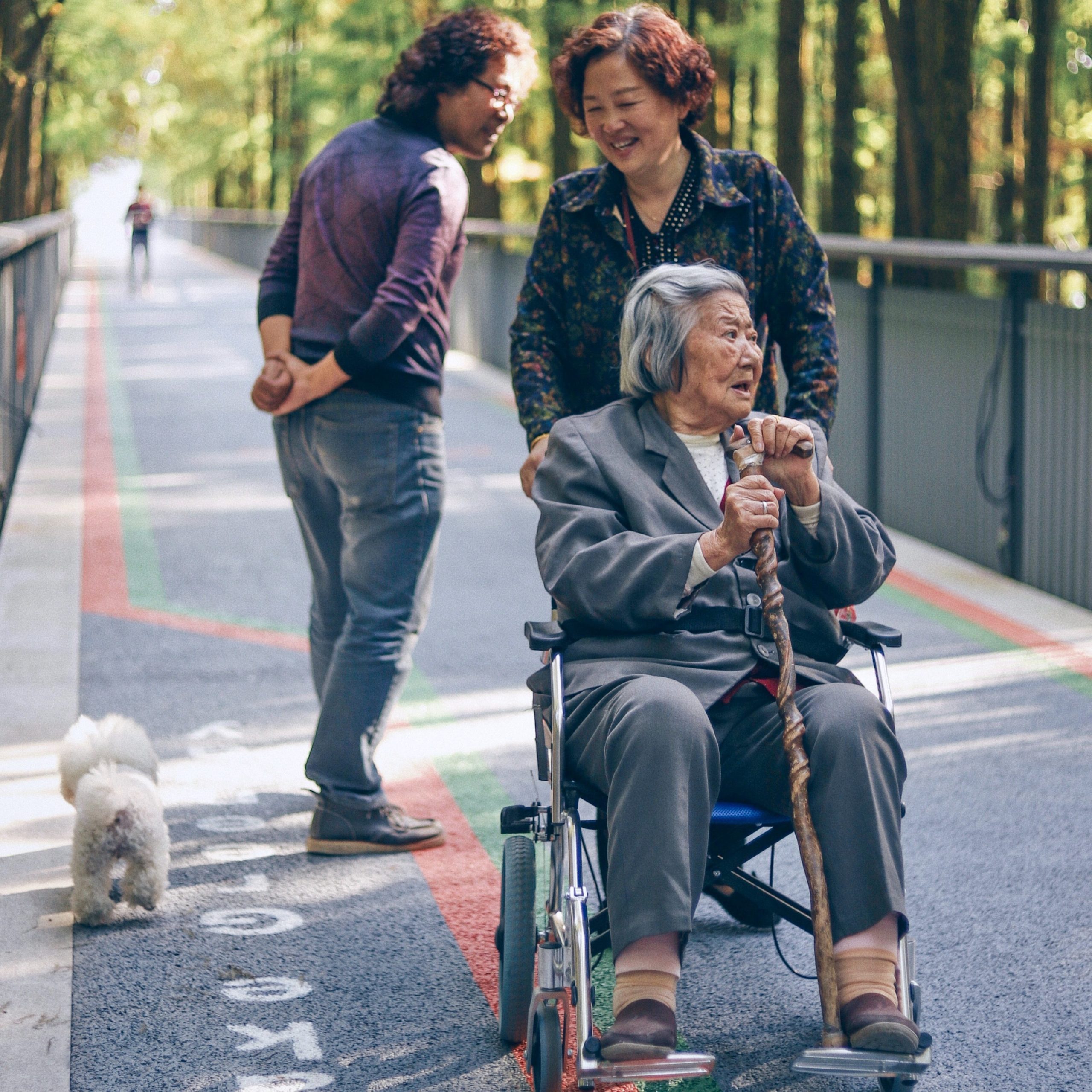 Asian American woman pushing her mother in a wheelchair along a walking bridge in woods. Man passing by them.