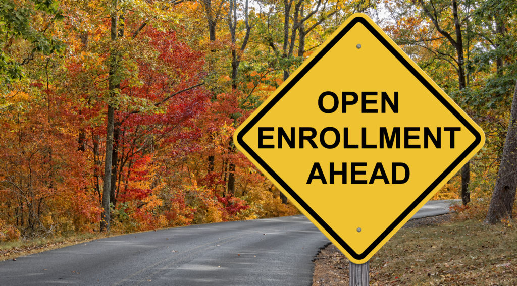 Open enrollment ahead road sign along road with colorful trees surrounding.