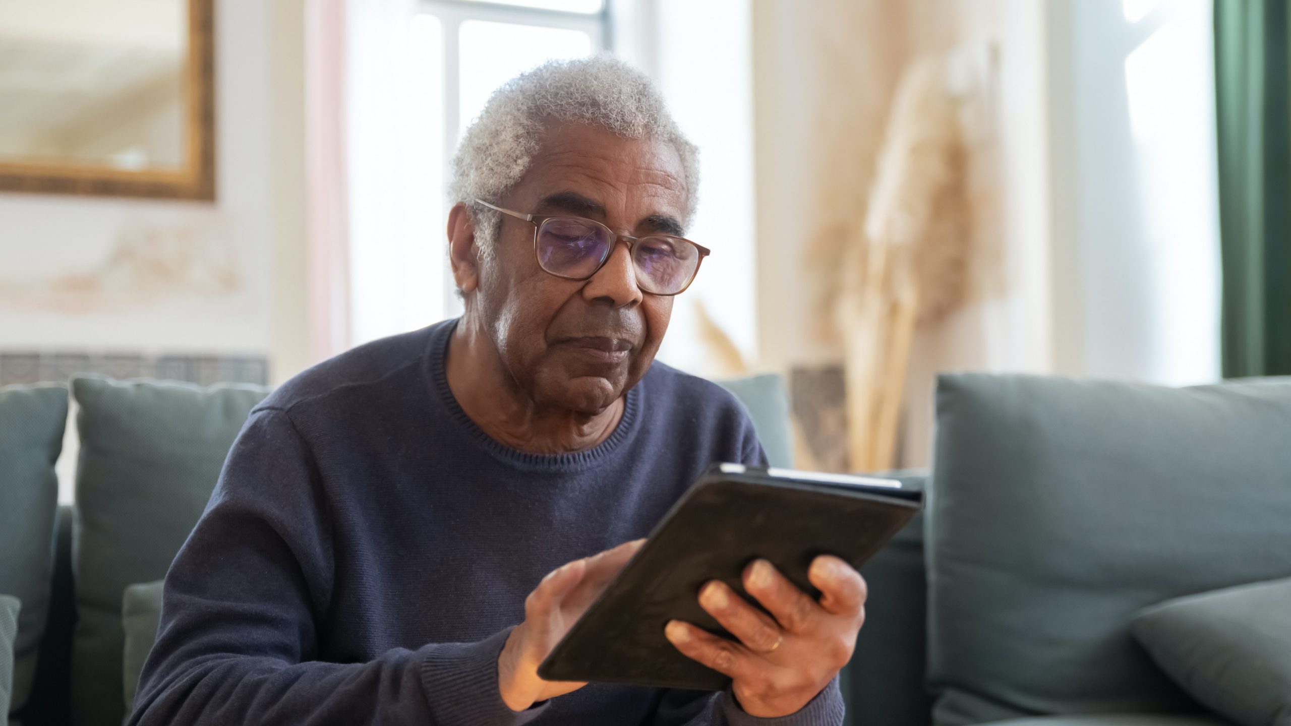 Man in glasses reading his tablet on a couch with a window behind him.