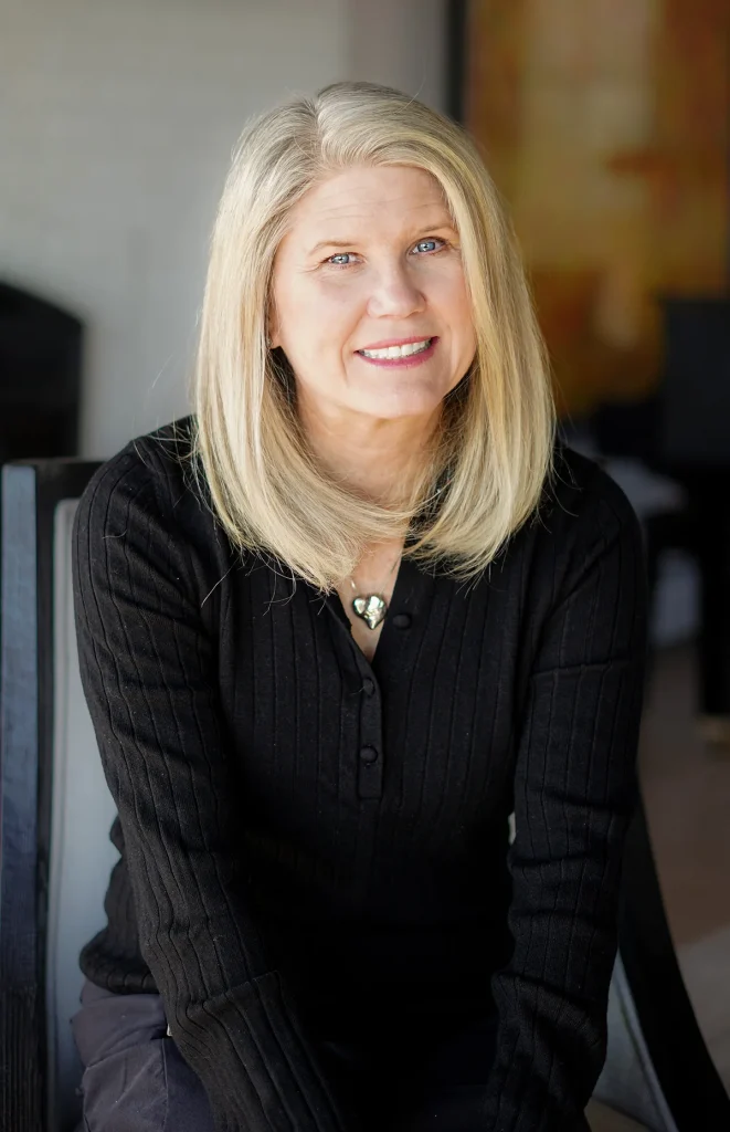 A smiling middle-aged woman with long blonde hair, wearing a black ribbed sweater, sits on a chair indoors with a blurred background.